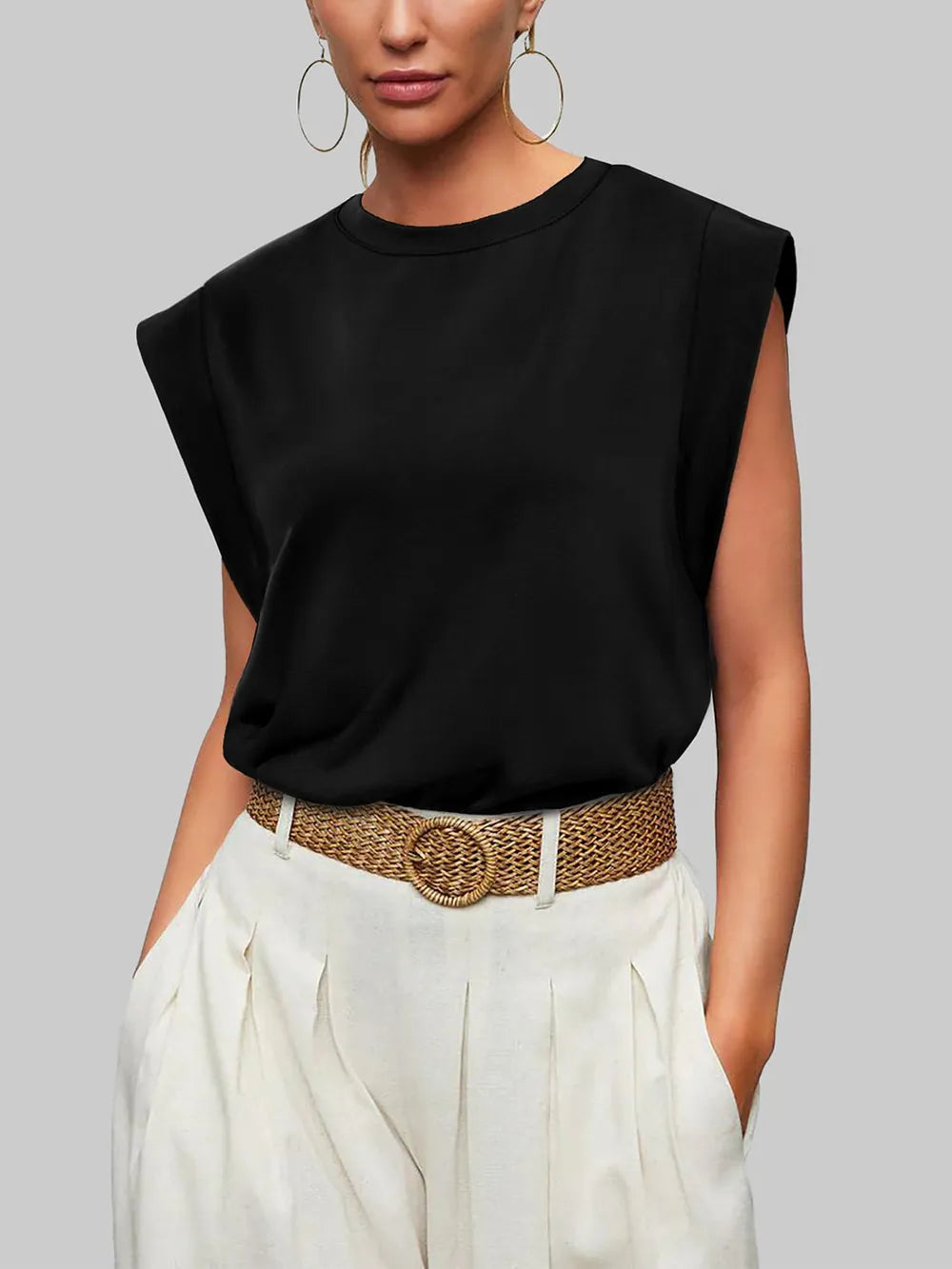 Woman wearing a black sleeveless top and white pants with a brown belt against a gray background