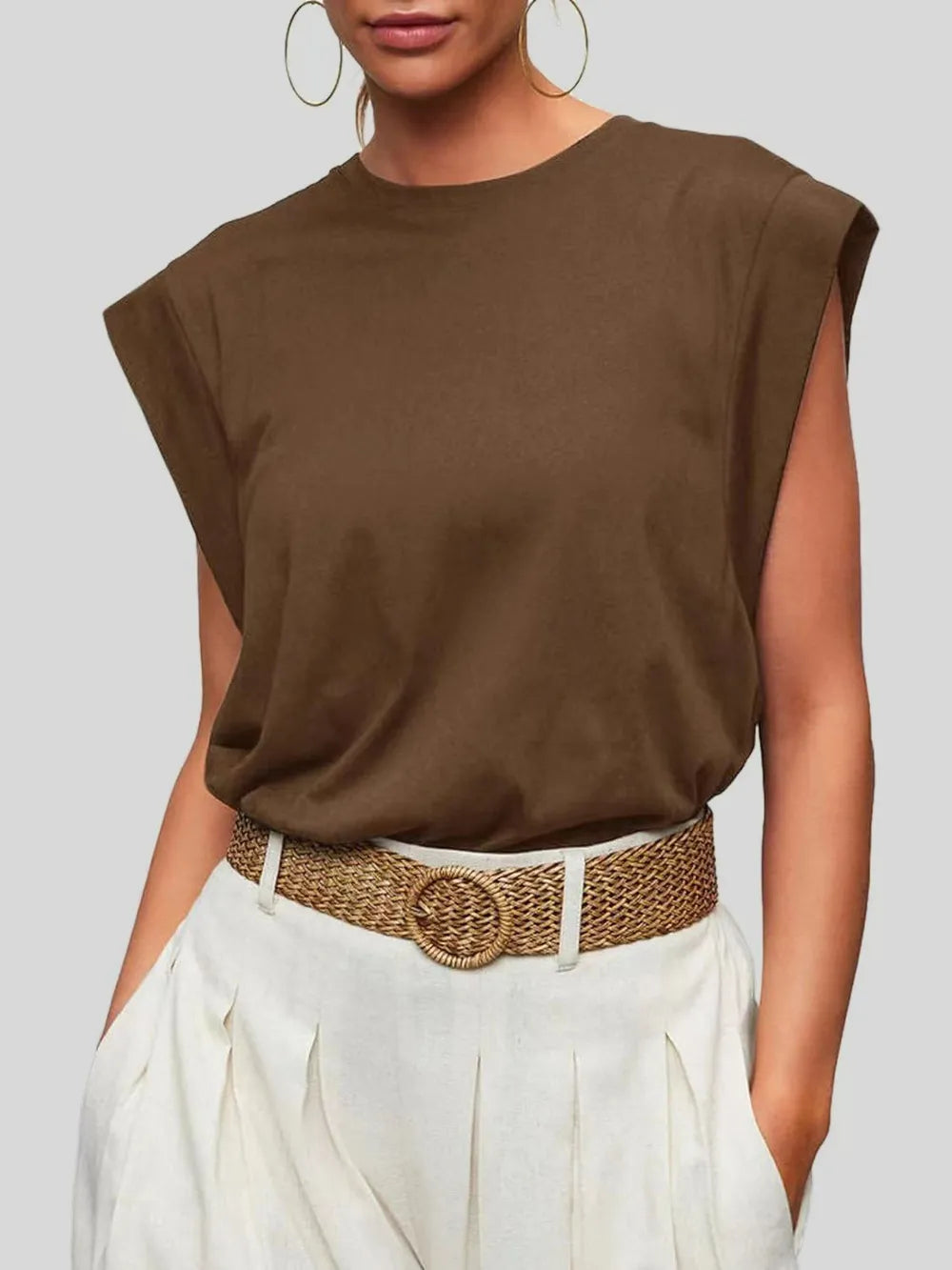 Person wearing a brown sleeveless top and white pants with a woven belt on a gray background