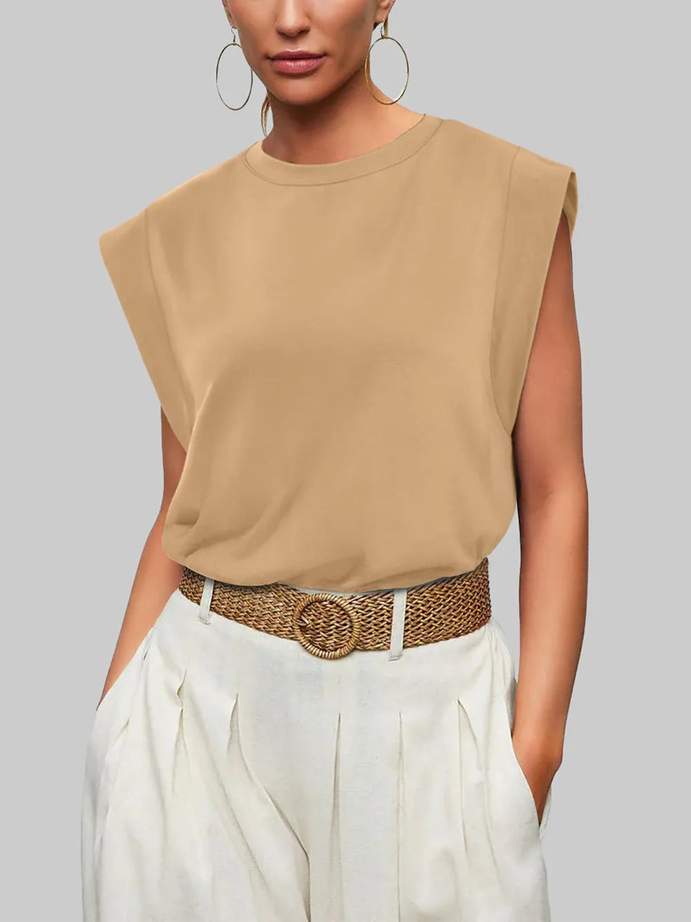 Woman wearing a beige sleeveless top and white pants with a brown belt against a gray background