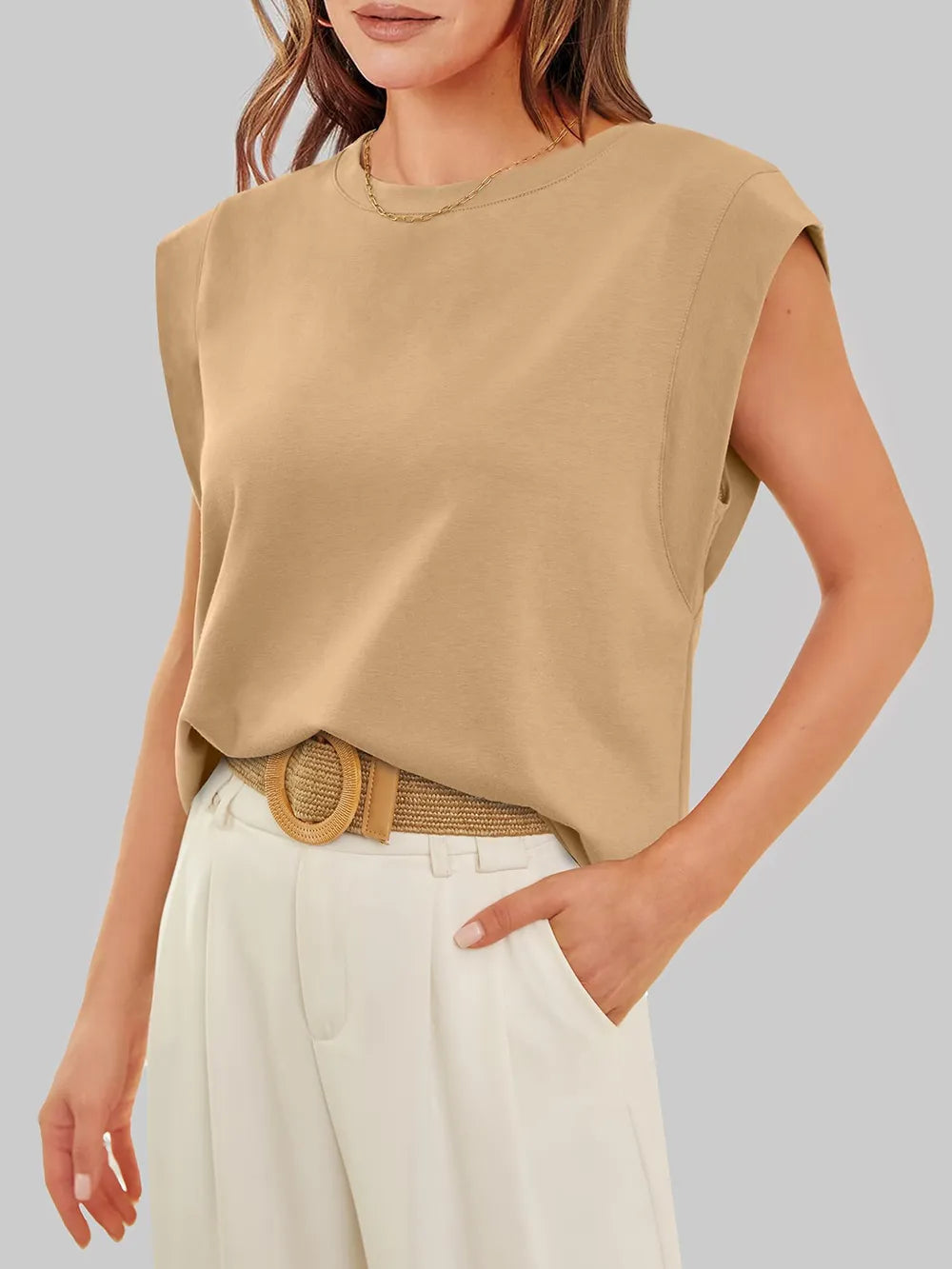 Woman wearing a beige sleeveless top and white pants against a gray background