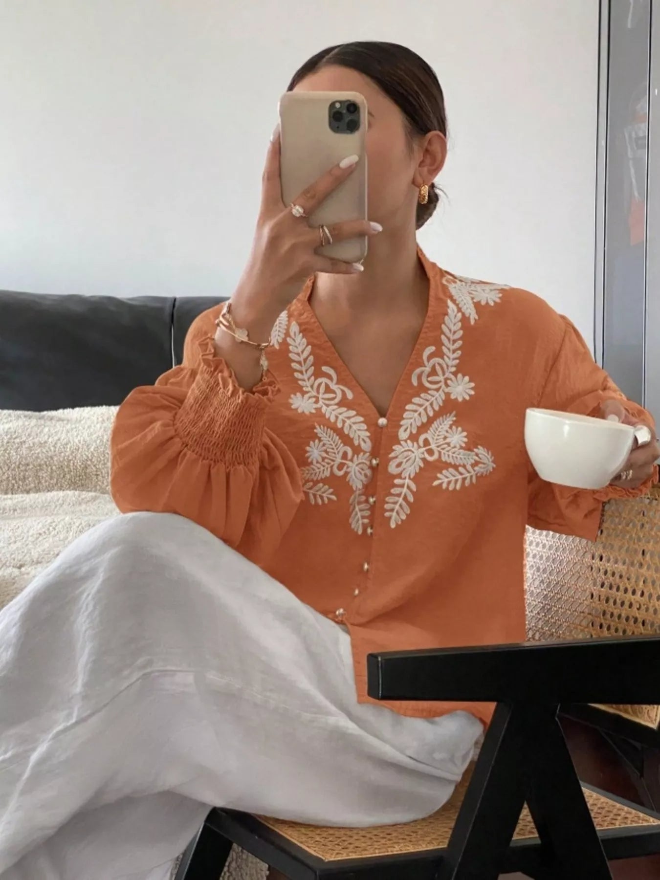 A woman sitting on a chair holding a white cup and smartphone in other hand wearing an orange embroidered button-front long-sleeve blouse 