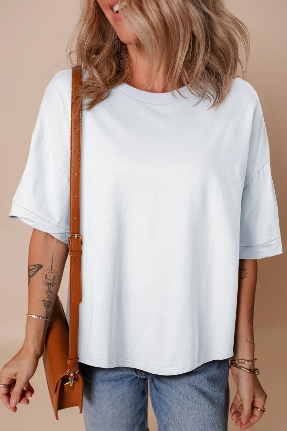 Person wearing a white t-shirt and blue jeans, holding a brown leather bag against a beige background