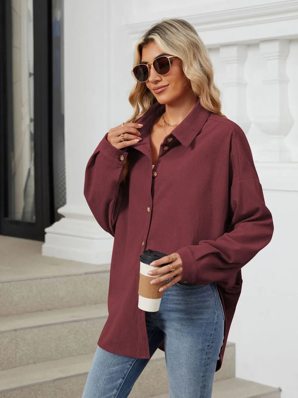 Woman wearing a burgundy shirt and sunglasses, holding a coffee cup, standing on steps.