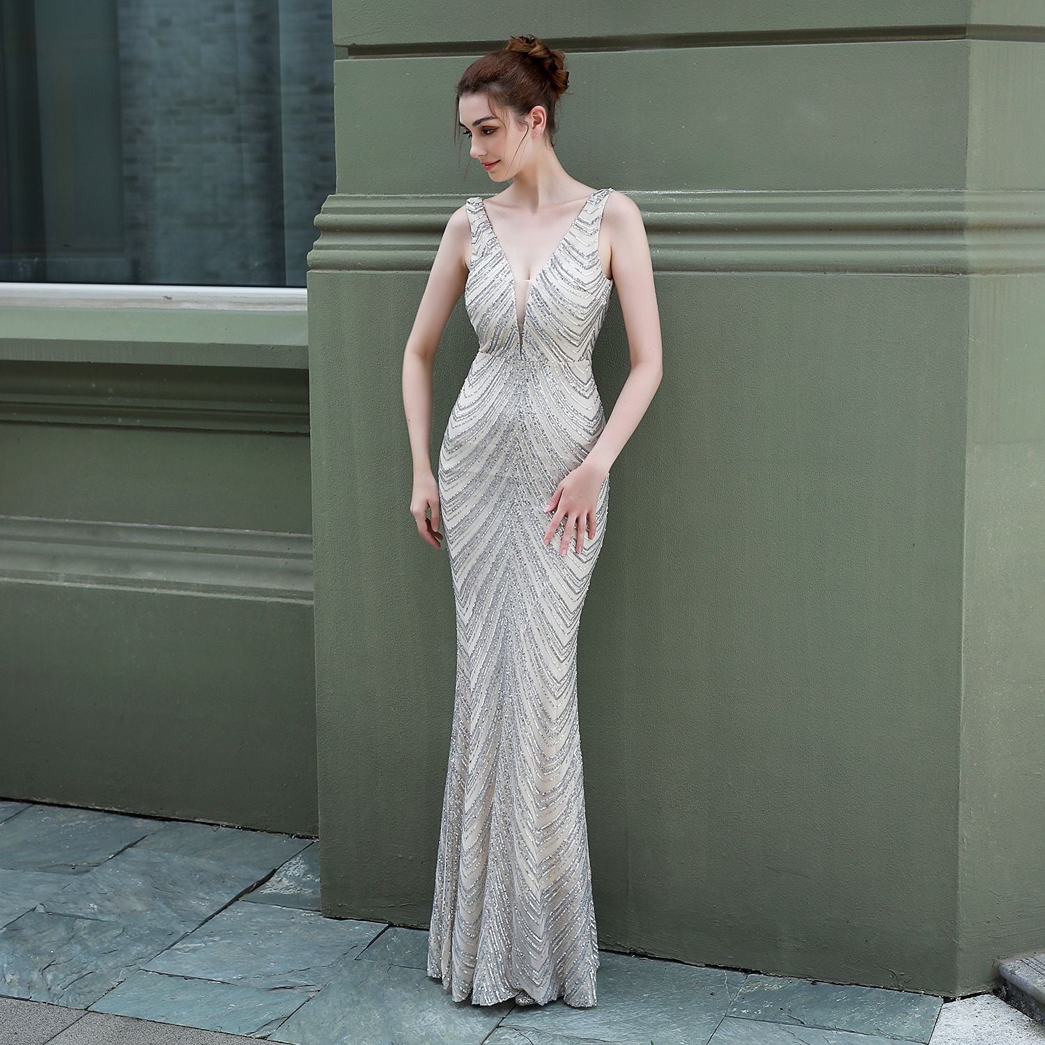 celeste-beaded-vneck-gown-white-front
Celeste white beaded V-neck evening gown with intricate sequin detailing, floor-length elegant maxi dress
Woman in a silver evening gown standing against a green wall.