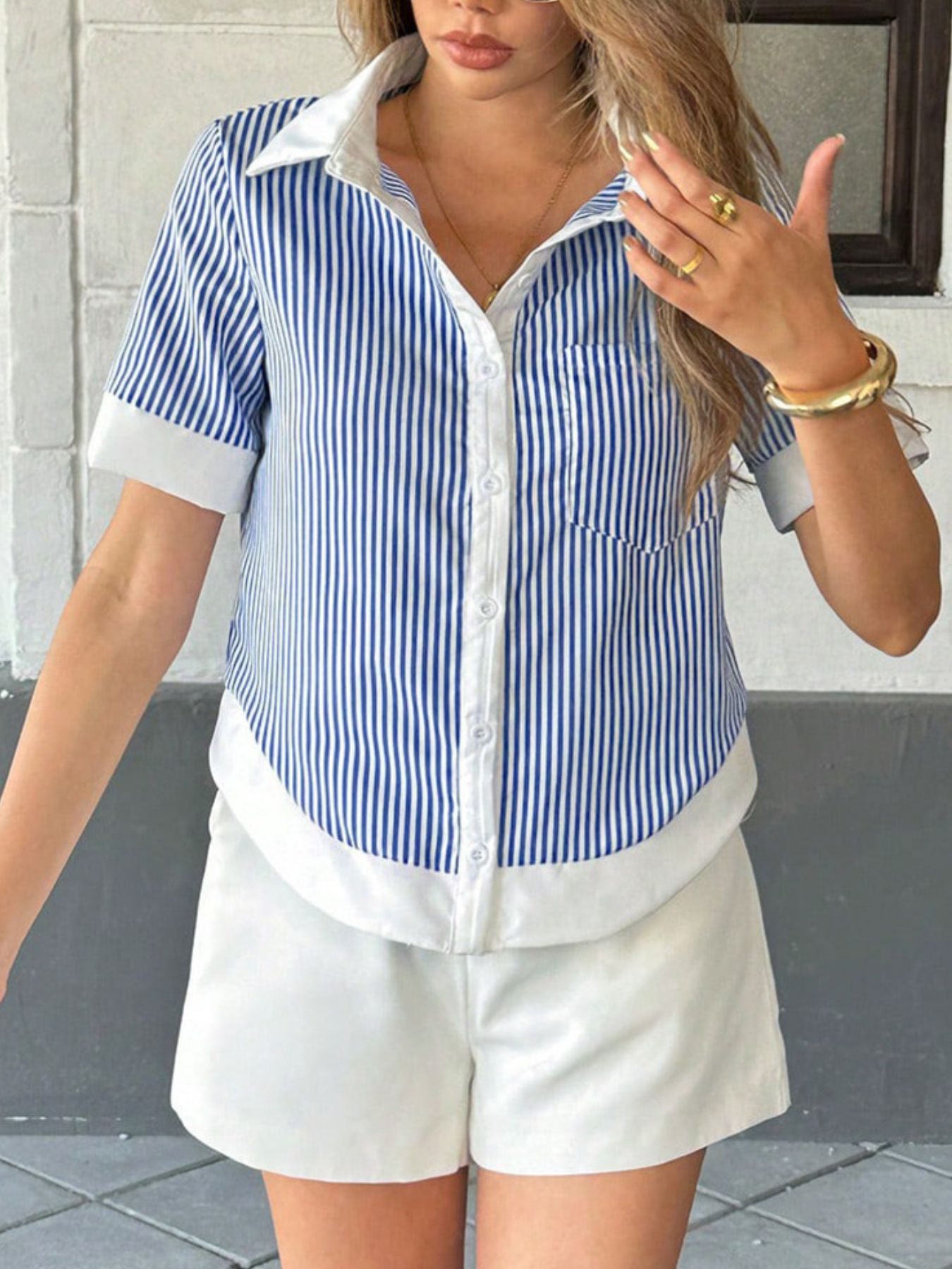 A woman wearing a blue and white striped shirt and white shorts, standing against a gray wall.
